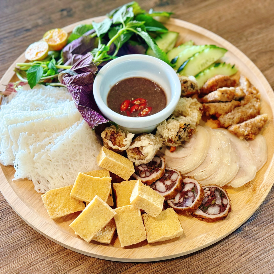 A wooden platter with various Vietnamese dishes including rice vermicelli, fried tofu, and sliced meats.