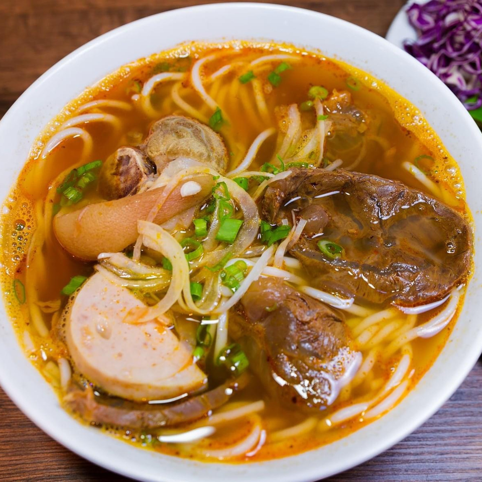 Close-up of a vibrant Vietnamese Bun Bo Hue soup with noodles, beef, pork, and herbs.